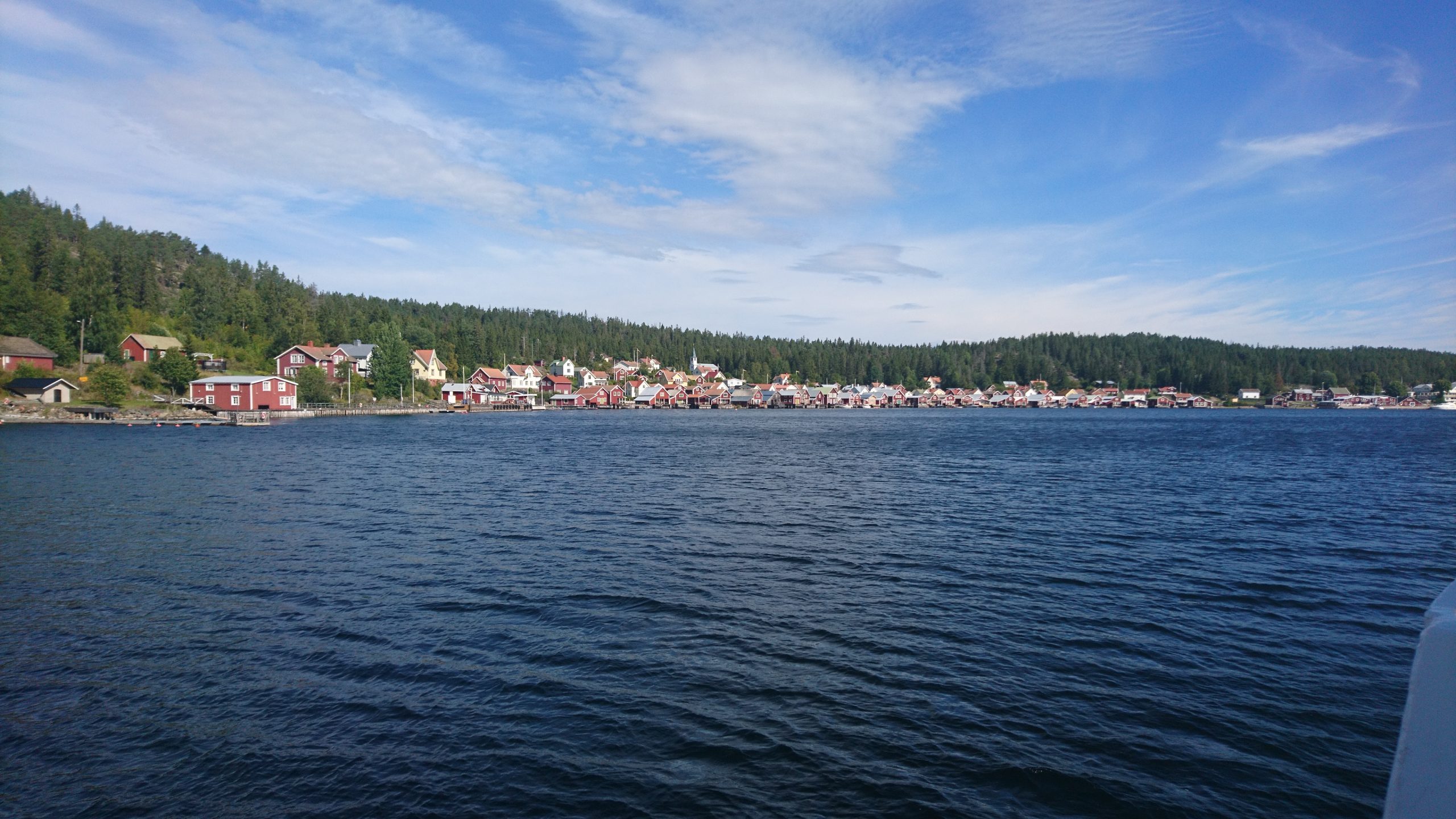 Röda sjöbodar och bostadshus i en lång rad längs vattnet i Ulvöhamn, med skogklädda berg bakom