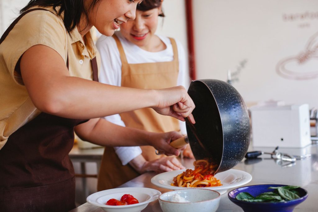 Två kursdeltagare i förkläden lägger upp pasta på tallrik under en matlagningskurs