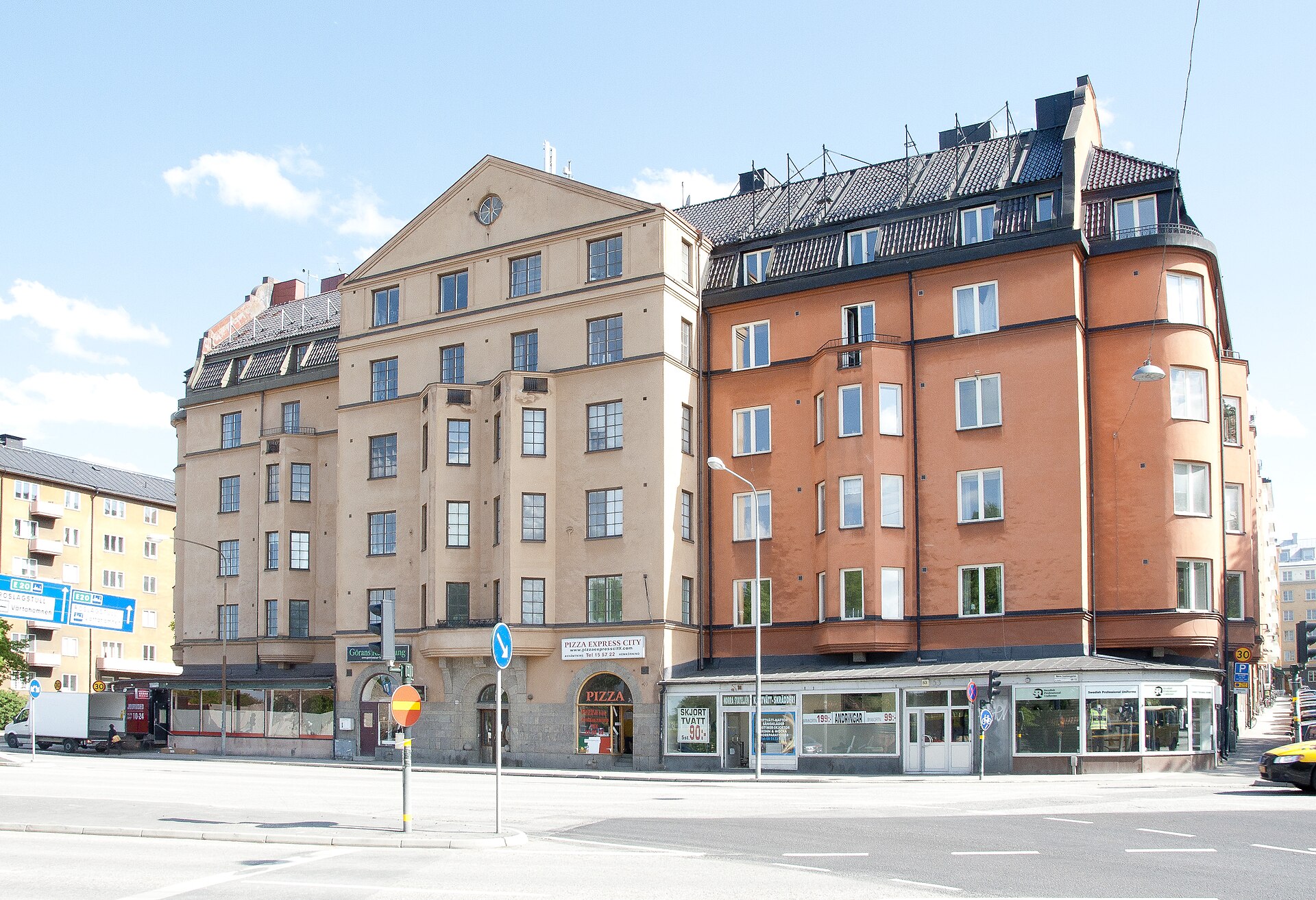 Två flerbostadshus i beige och rödorange puts med butiker i bottenvåningen i ett gathörn i Stockholm