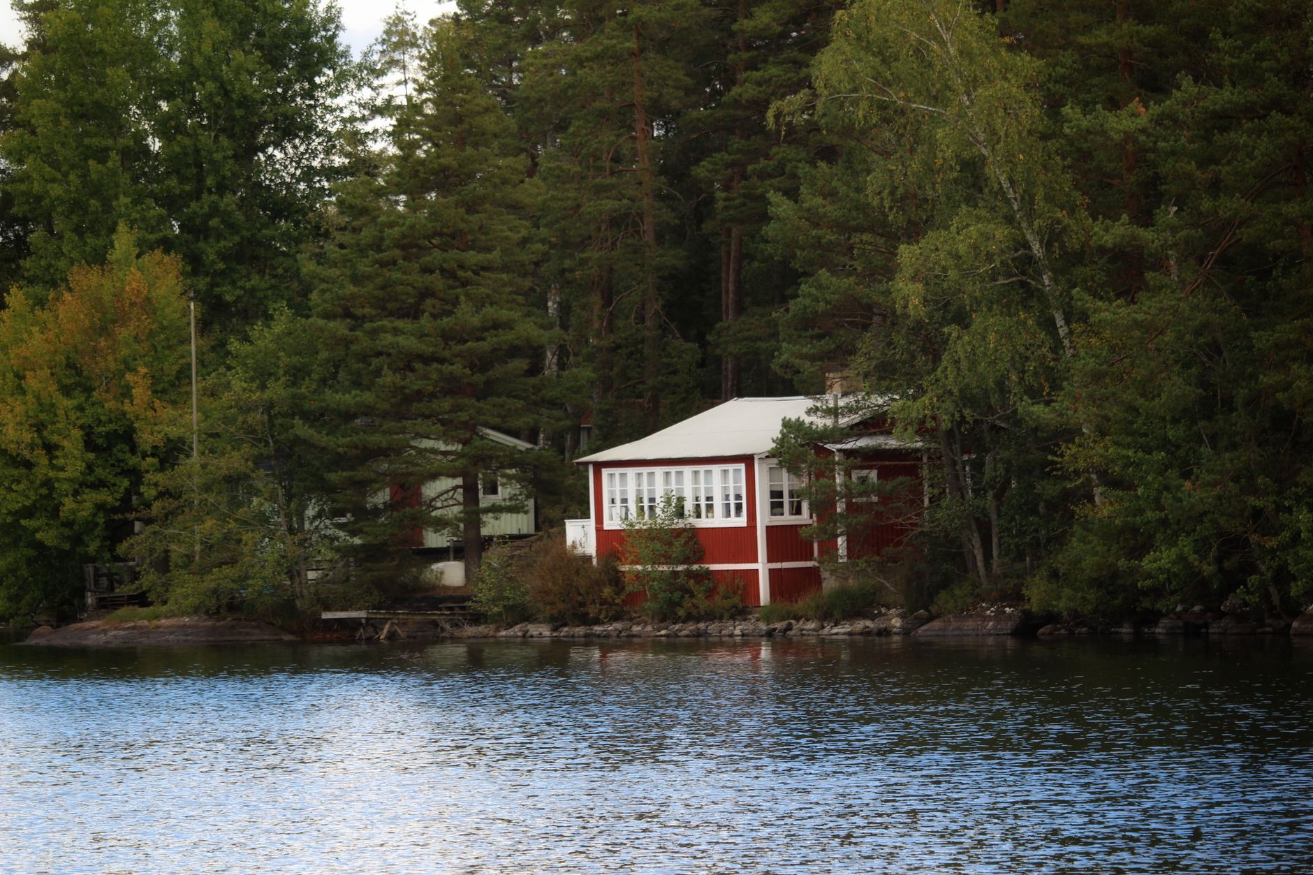 Röd stuga med inglasad veranda vid en sjö, omgiven av tallar och björkar