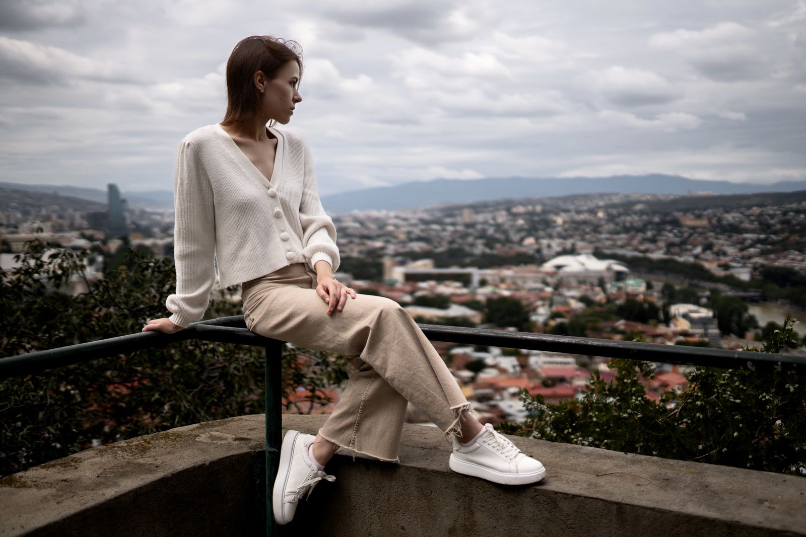 Kvinna i beige byxor och kofta med vita lädersneakers på en stadsbalkong