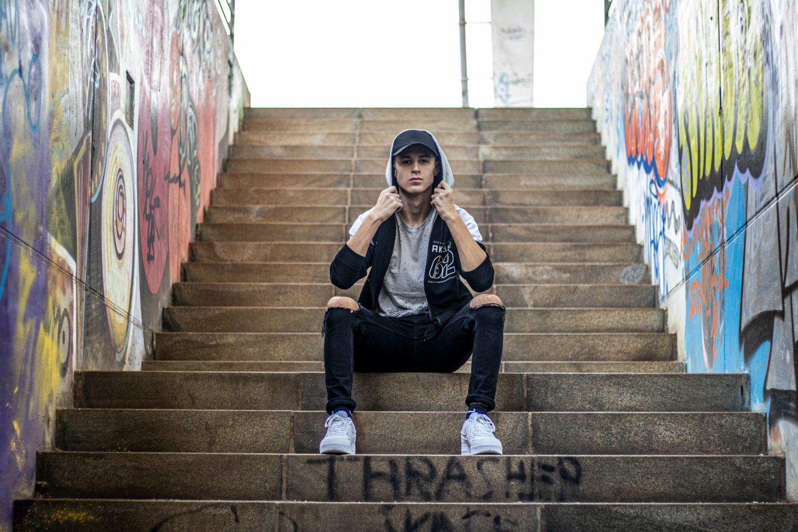 Ung man i keps, huvtröja och slitna jeans i en graffitiklädd trappa med vita sneakers