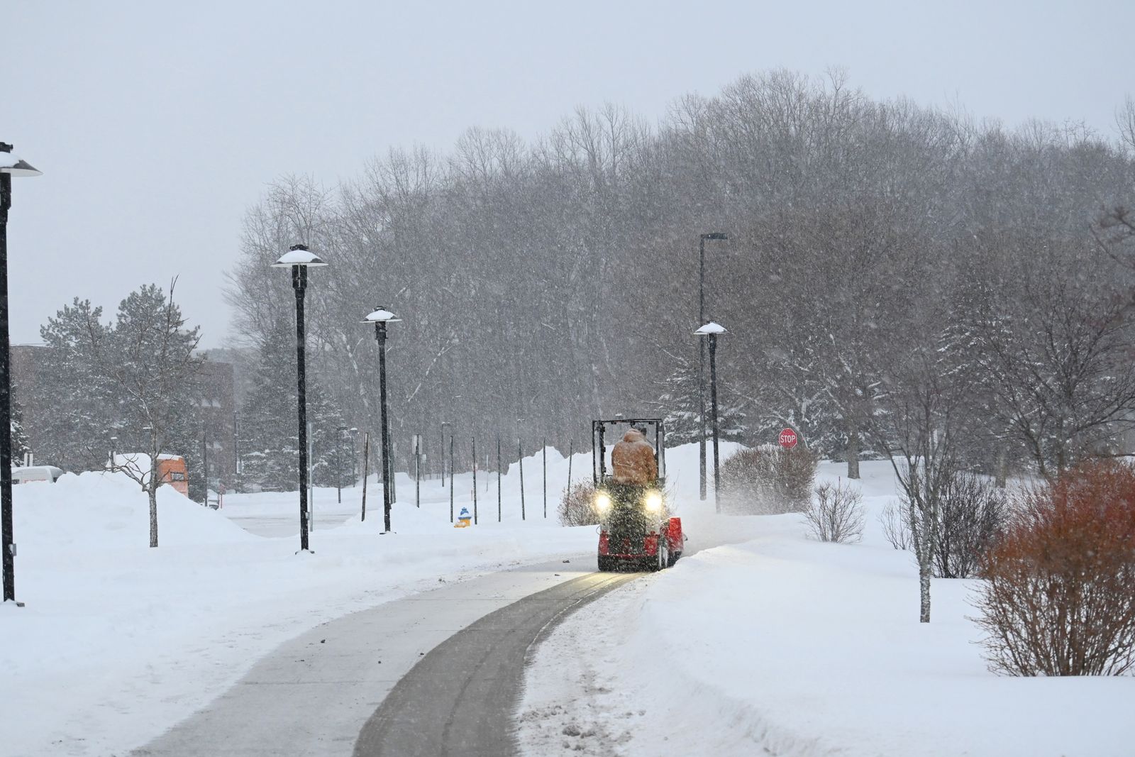 Kompakt snöröjningsmaskin plogar en gångväg under snöfall, med lyktstolpar och snötäckta träd längs vägen