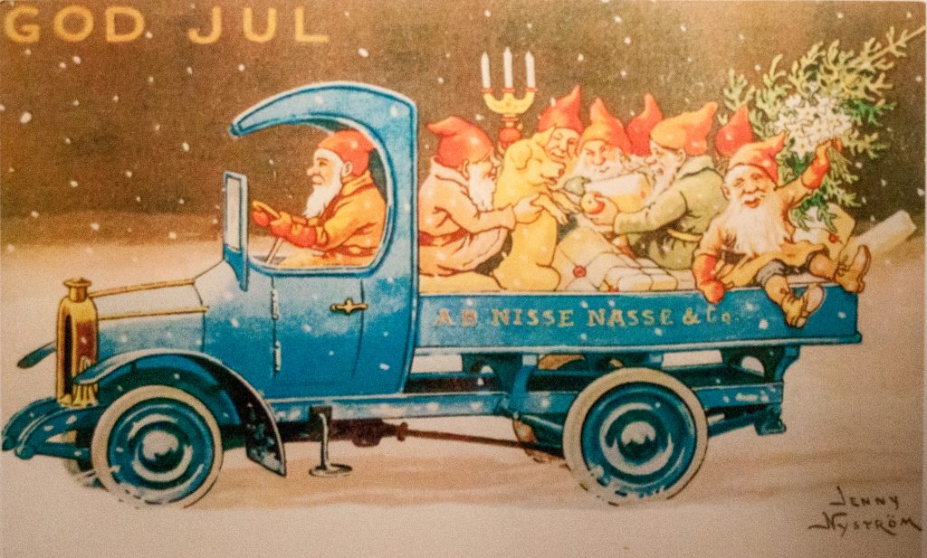 Julkort av Jenny Nyström där rödmössade tomtar åker lastbil med granris och julklappar genom snöfall