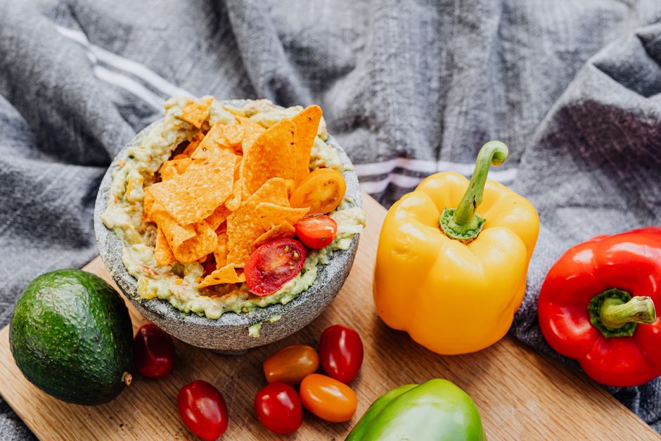 Skål med guacamole och nachos, omgiven av avokado, tomat och paprika