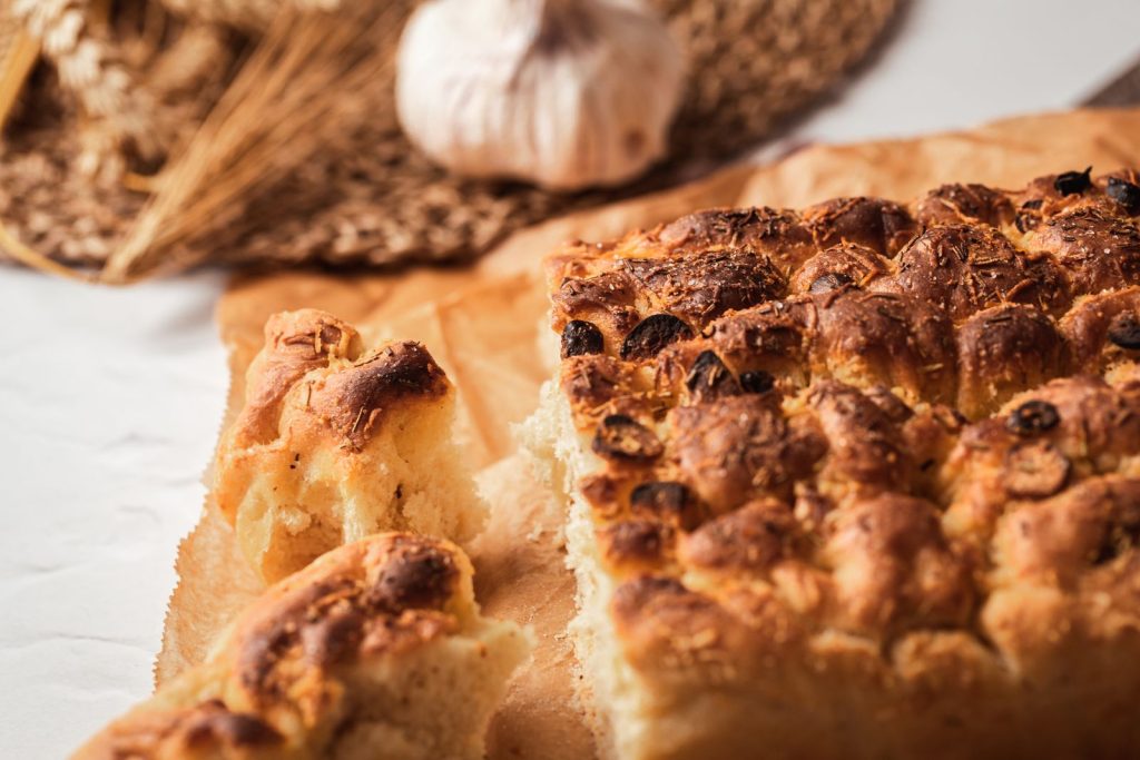 Nybakad focaccia med rosmarin och oliver, uppbruten i bitar på bakplåtspapper