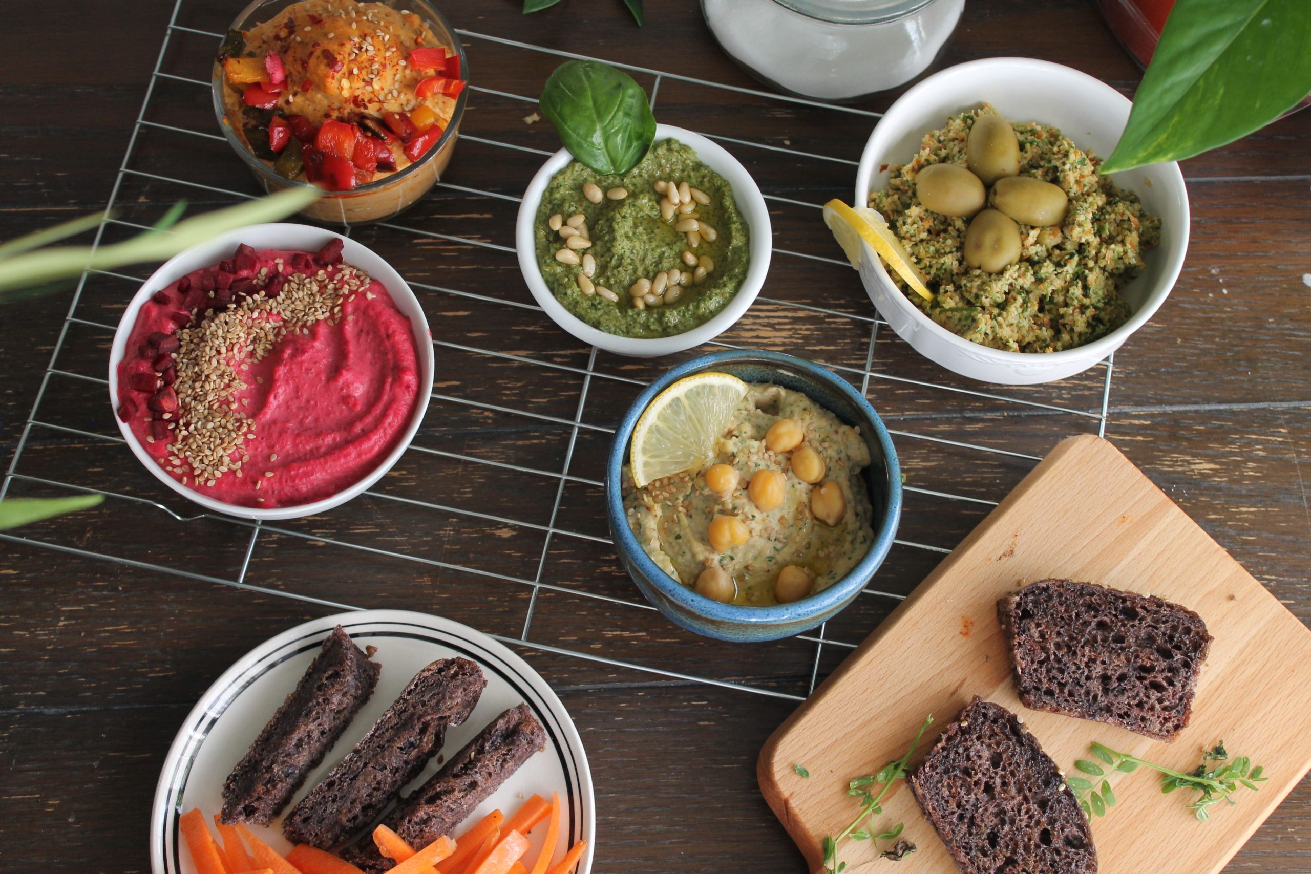 Fem skålar med veganska röror, mörkt rågbröd och morotsstavar på ett träbord