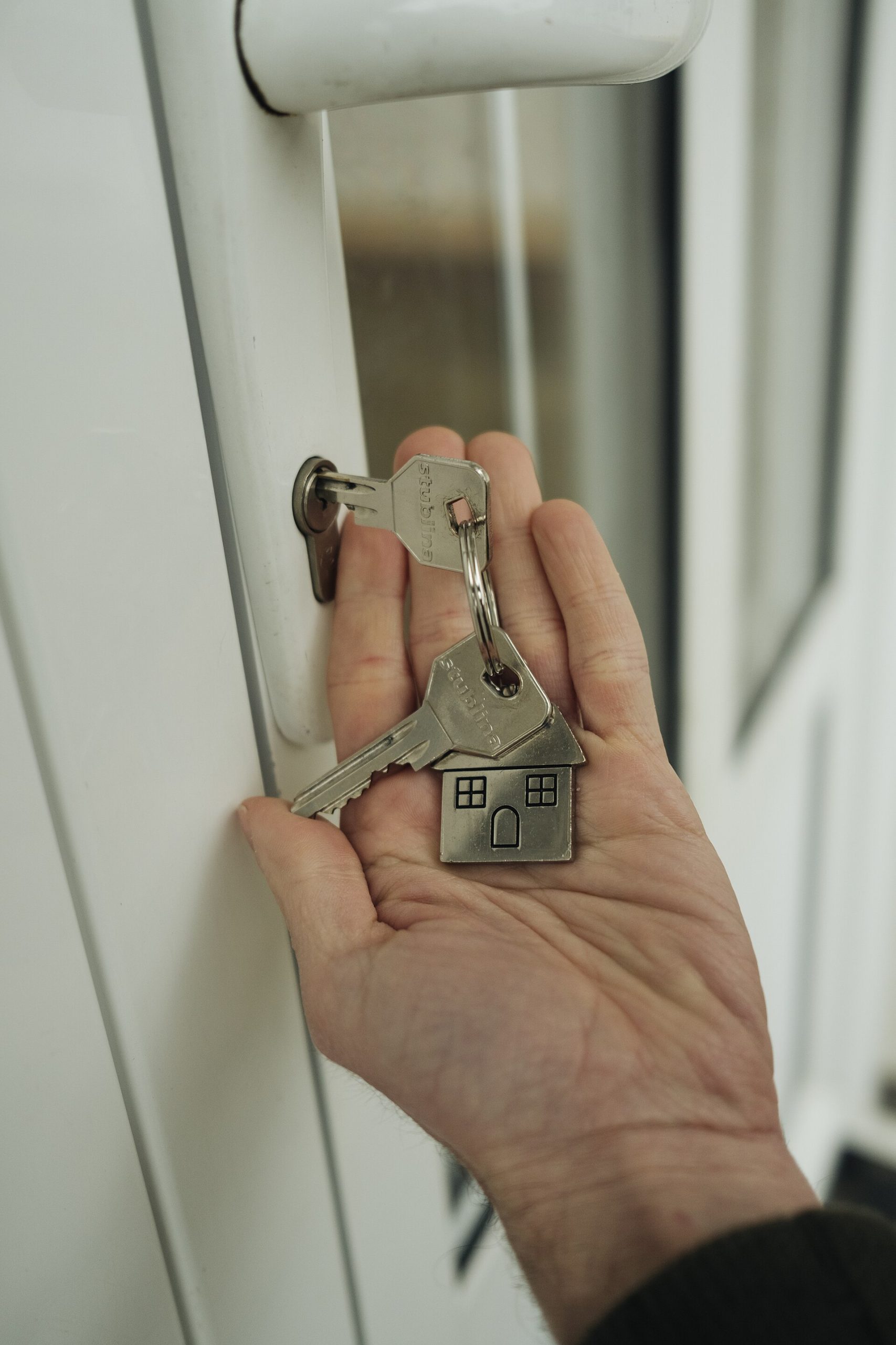 En hand håller ett nyckelknippe med en husformad bricka medan nyckeln sitter i låset på en vit ytterdörr.