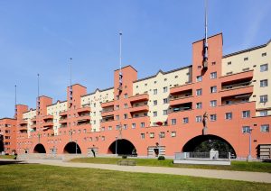 Karl-Marx-Hofs långa fasad i terrakotta och gult med valvbågar och flaggstångstorn, med gräsmatta framför.