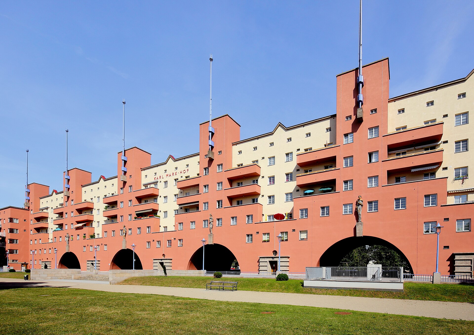 Karl-Marx-Hofs långa fasad i terrakotta och gult med valvbågar och flaggstångstorn, med gräsmatta framför.