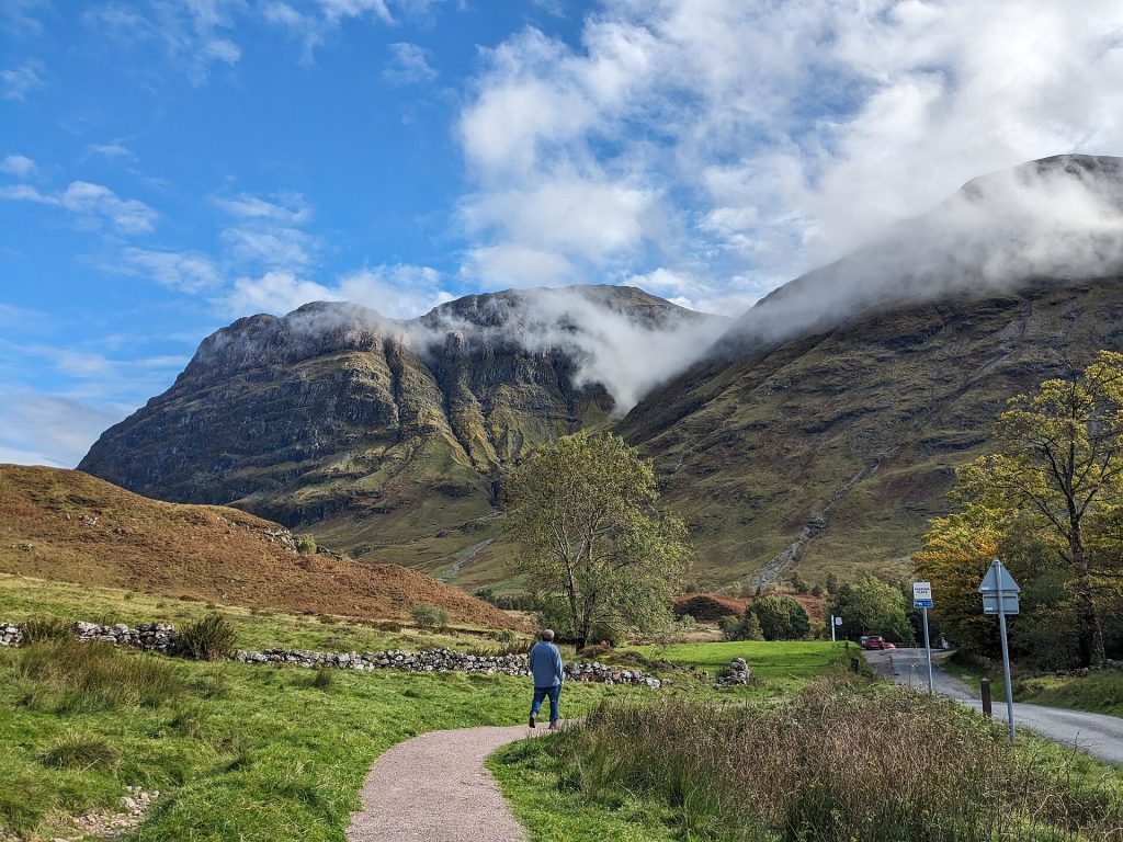 Vandrare på grusstig vid gamla vägen genom Glencoe med bergssidan och en skylt för mötesplats i bakgrunden