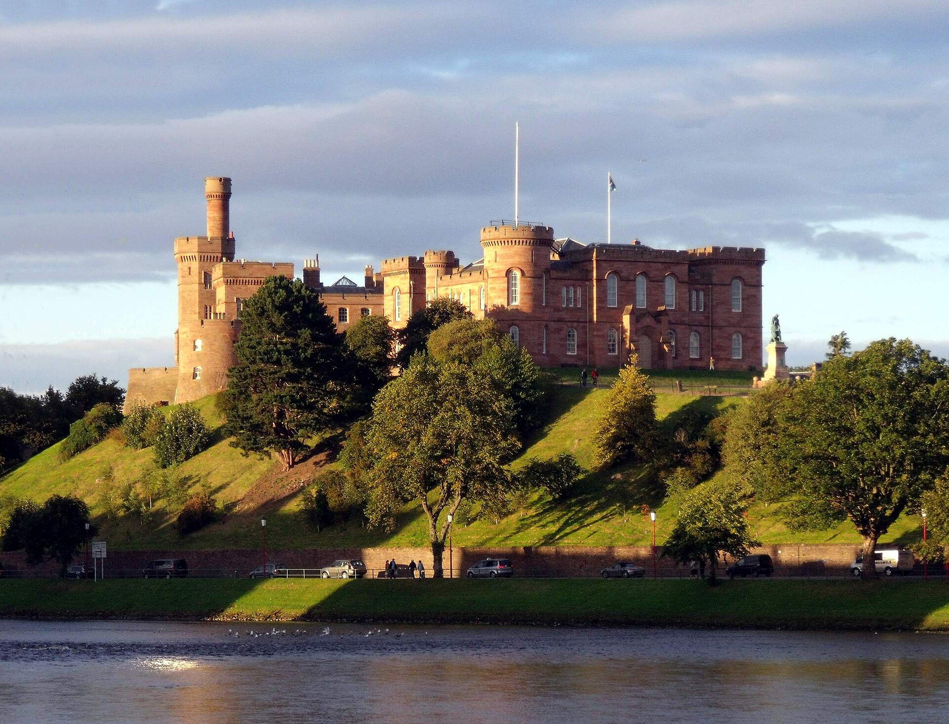 Inverness Castle i rödbrun sandsten på kullen ovanför floden Ness, i kvällssol med träd på slänten ned mot vattnet