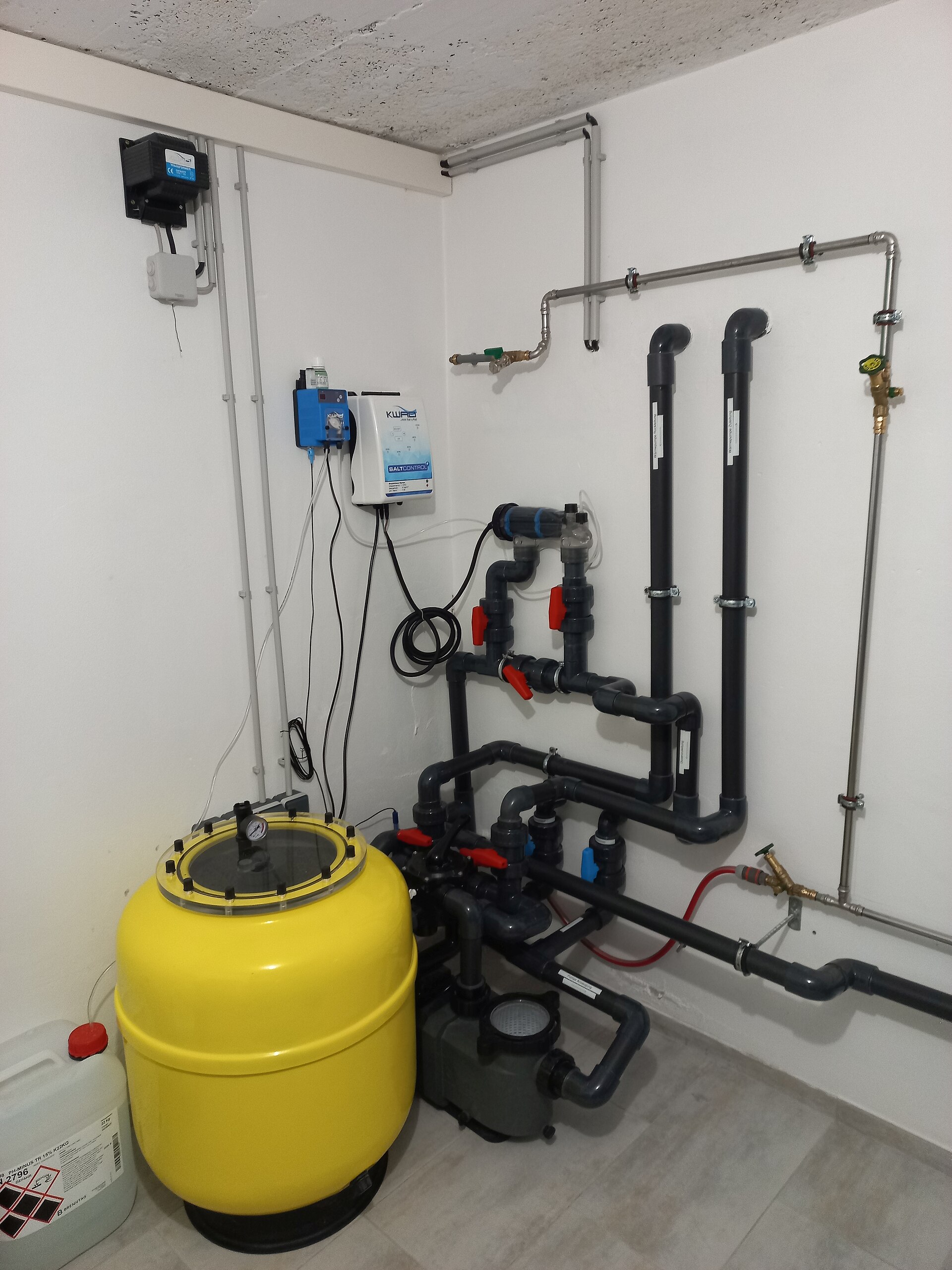 Gul sandfiltertank med manometer, pump och rör med ventiler i ett poolteknikrum