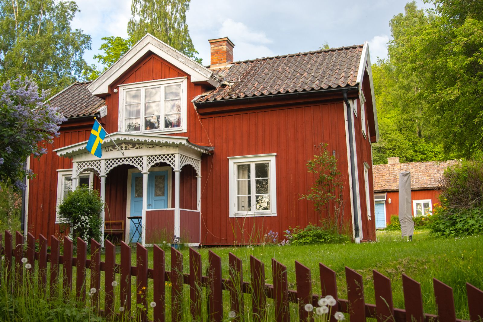 Röd sommarstuga med vit veranda, syrener i blom och svensk flagga