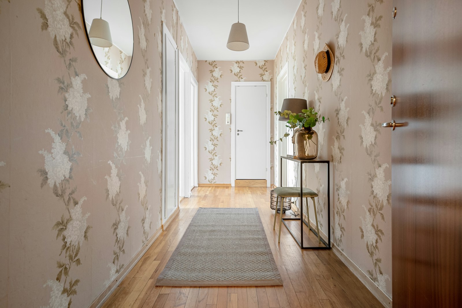 Smal hall med blekrosa blomtapet på båda väggarna, rund spegel och trasmatta på parkett