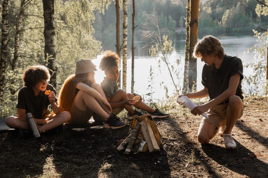 Fyra ungdomar sitter vid en lägereld vid en sjö i kvällssol