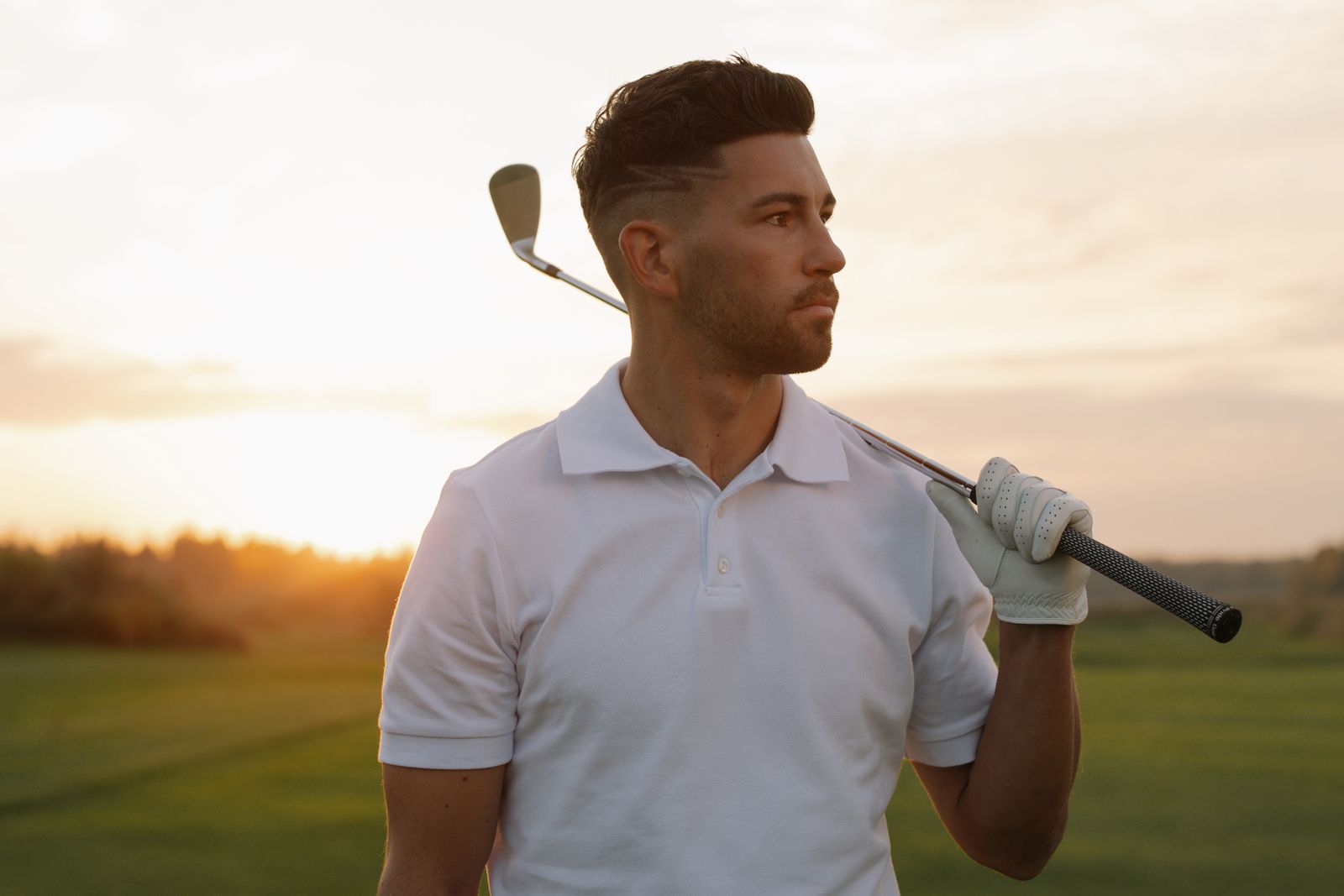 Stilfull golfare i vit piké med klubba över axeln i solnedgång på banan