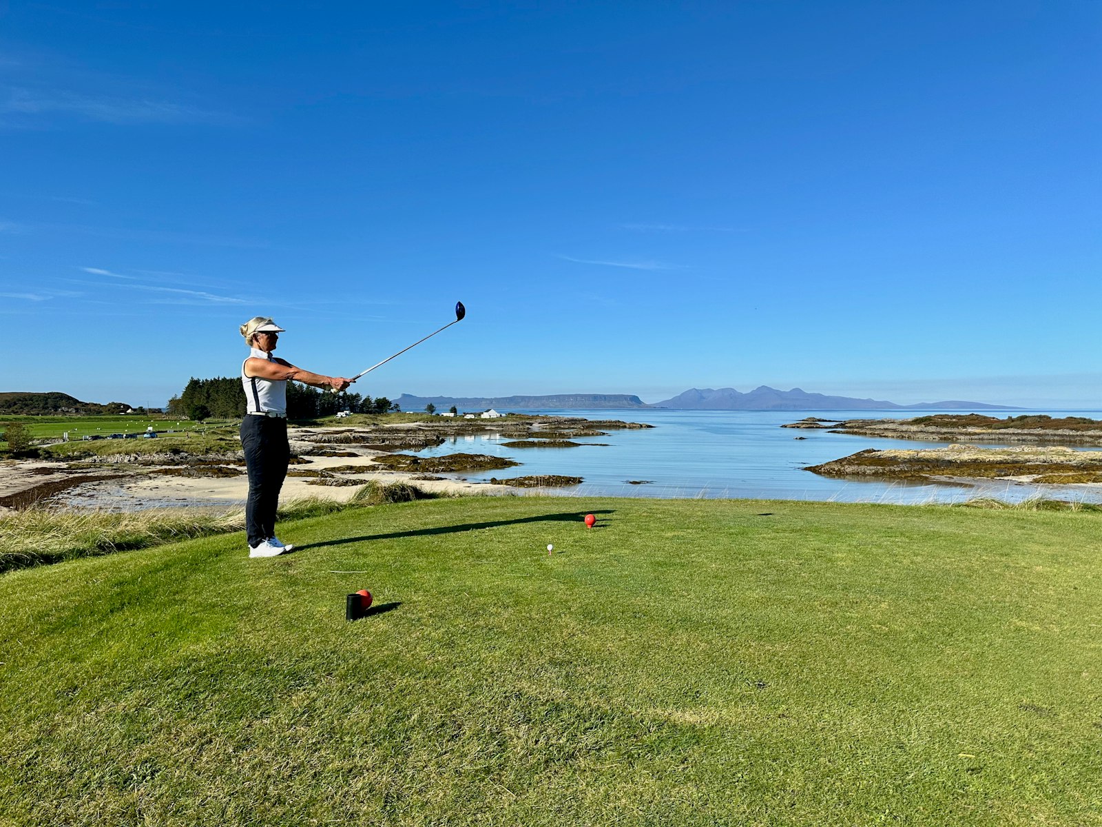 Golfare står kvar i utslagsställningen efter ett slag från en tee vid havet, med röda teemarkeringar i gräset