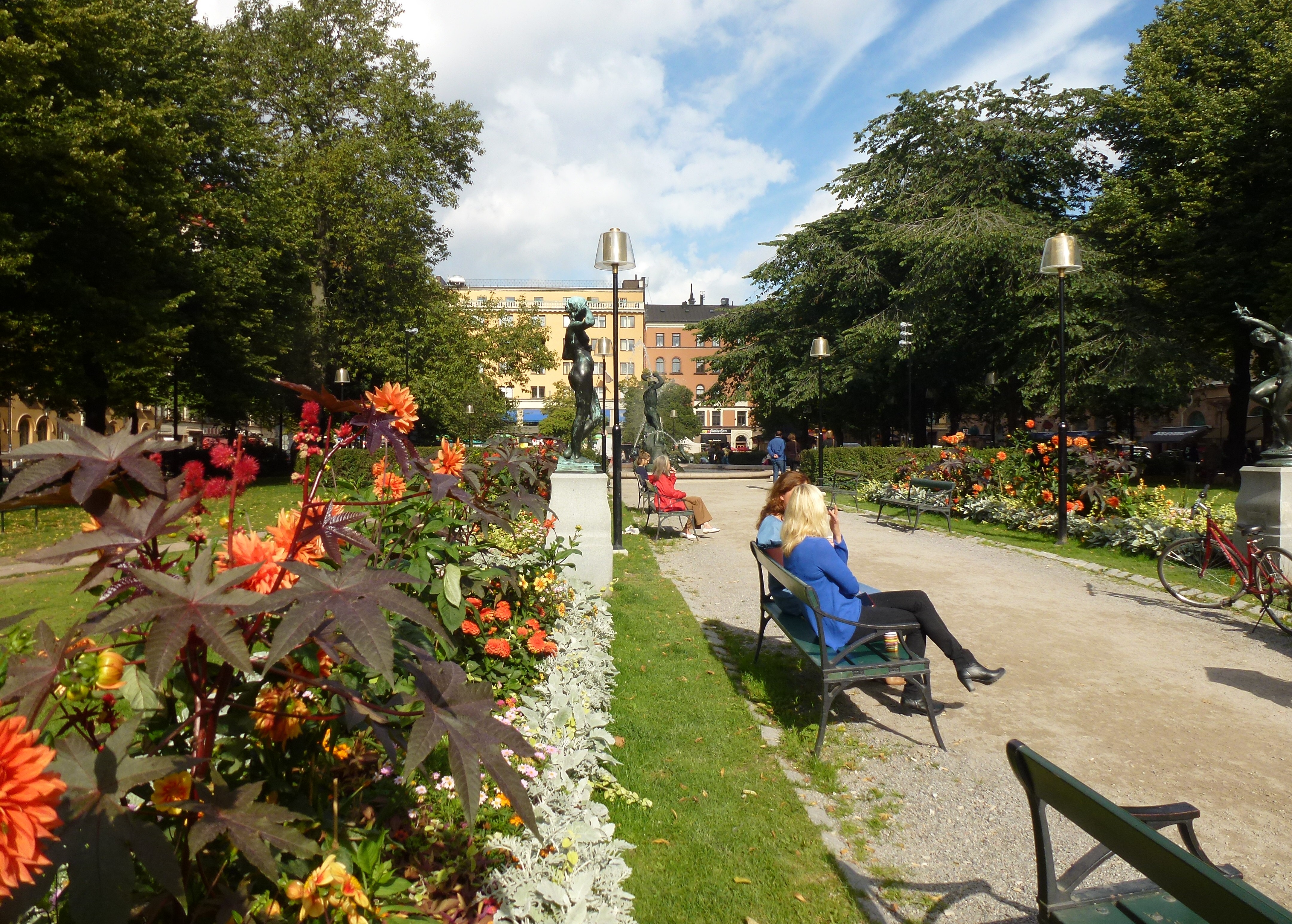 Blomsterrabatter, grusgång och parkbänkar på Mariatorget med fontänen Tors fiske i bakgrunden