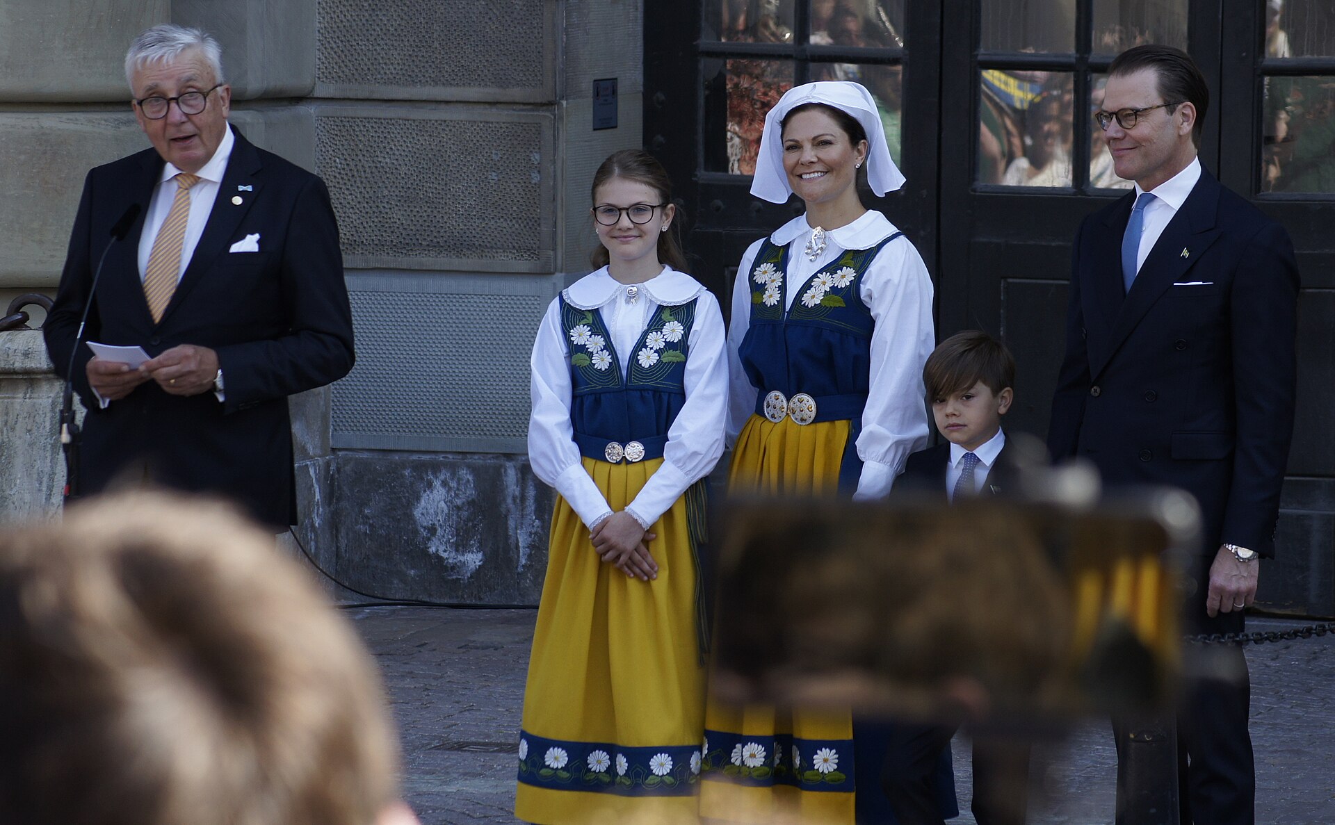 Kronprinsessan Victoria och prinsessan Estelle i folkdräkt tillsammans med prins Oscar och prins Daniel utanför slottet.
