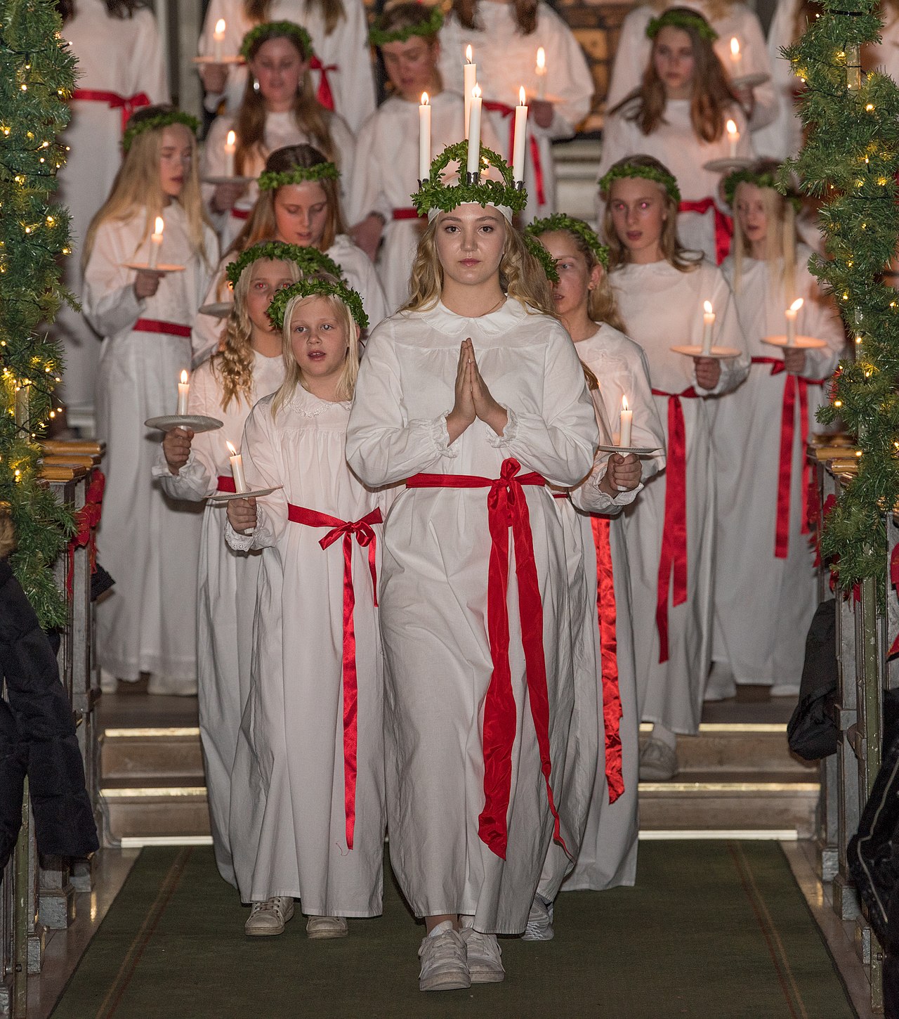 Lucia med ljuskrona och tärnor i vita klädnader med levande ljus i ett luciatåg