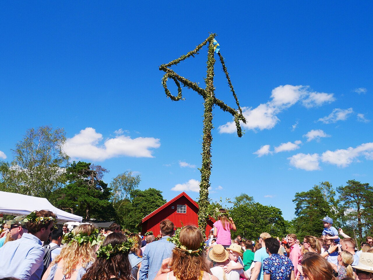 Svensk midsommarstång klädd med löv och blommor, människor med blomsterkransar och röd stuga