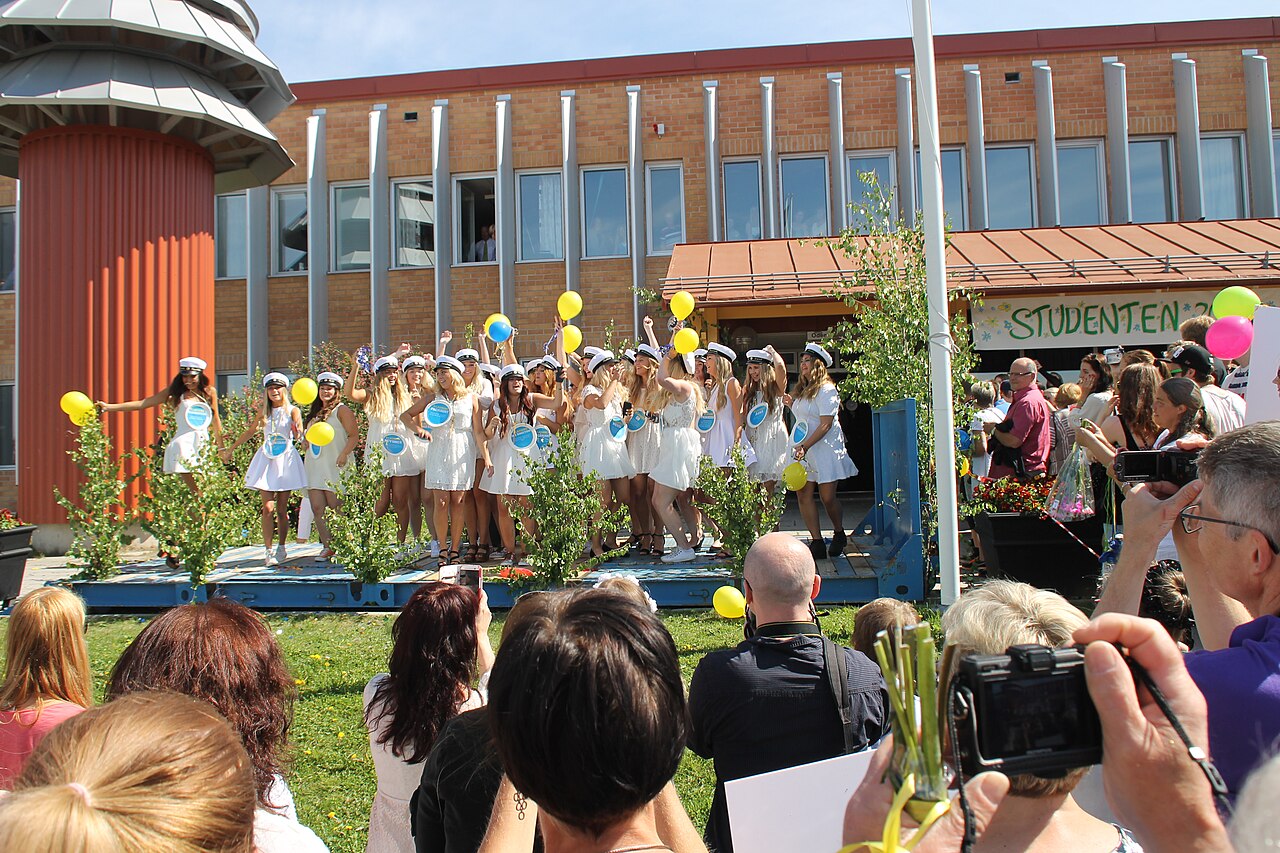 Studenter i vita mössor firar på ett flak med blå och gula ballonger framför en jublande publik