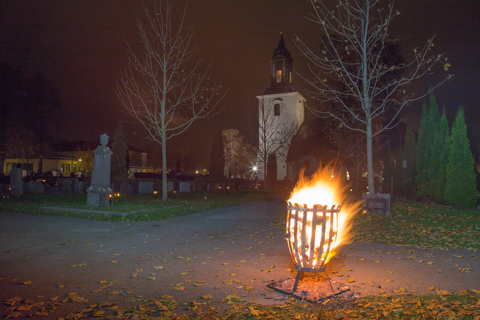 Kyrkogården vid Hedemora kyrka en mörk kväll på alla helgons dag, med en eldkorg på gången och tända gravljus mellan gravstenarna.