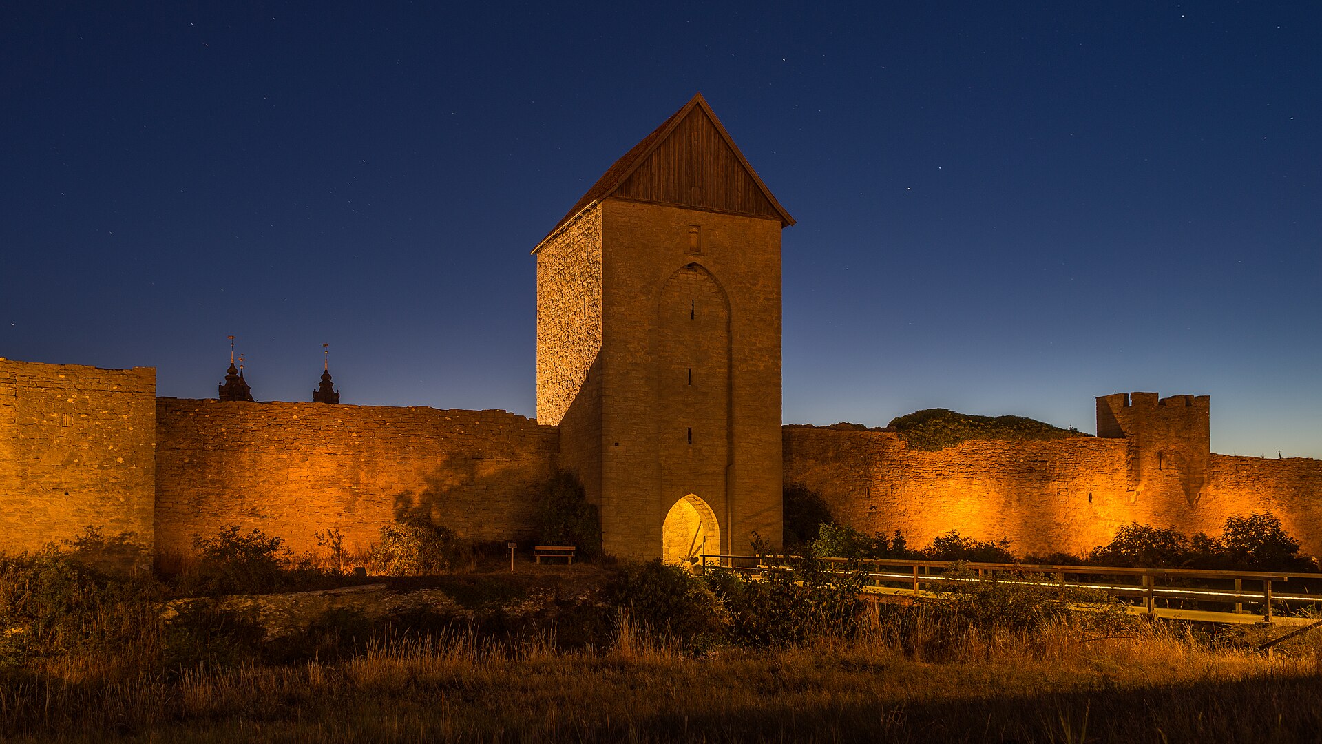 The medieval ring wall of Visby on Gotland lit up at dusk, with a tall stone gate tower and the cathedral spires behind the wall