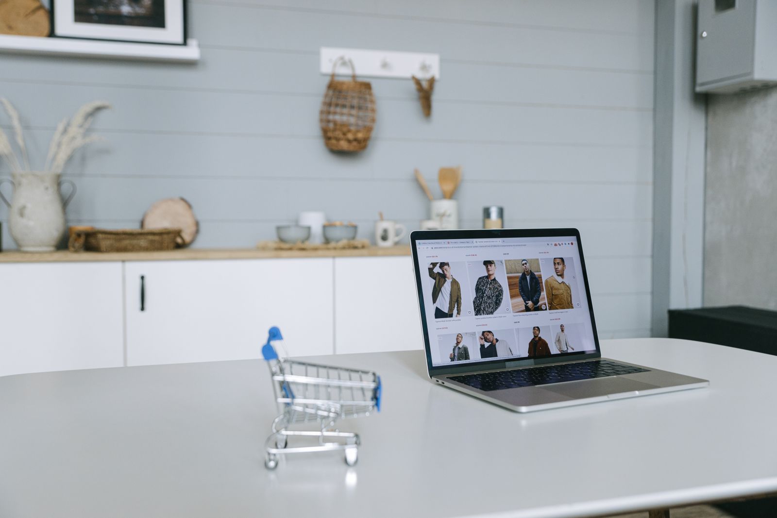 Bärbar dator som visar produktlistan i en webbutik, med en liten kundvagn bredvid på ett ljust köksbord