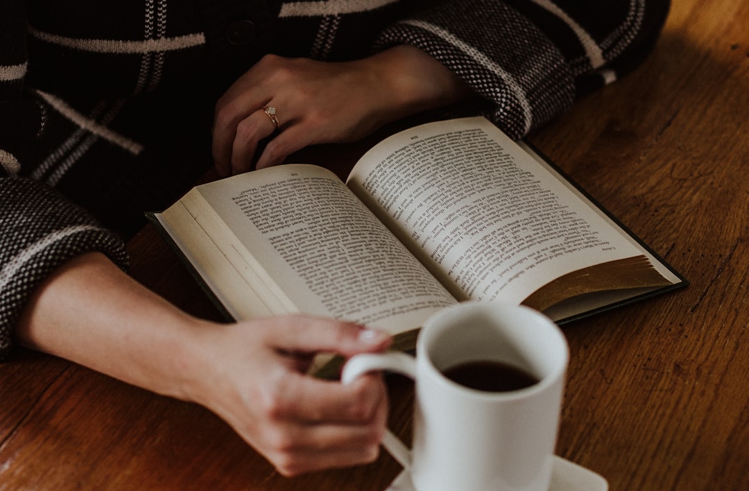 Händer som håller en kaffekopp vid en uppslagen bok på ett träbord