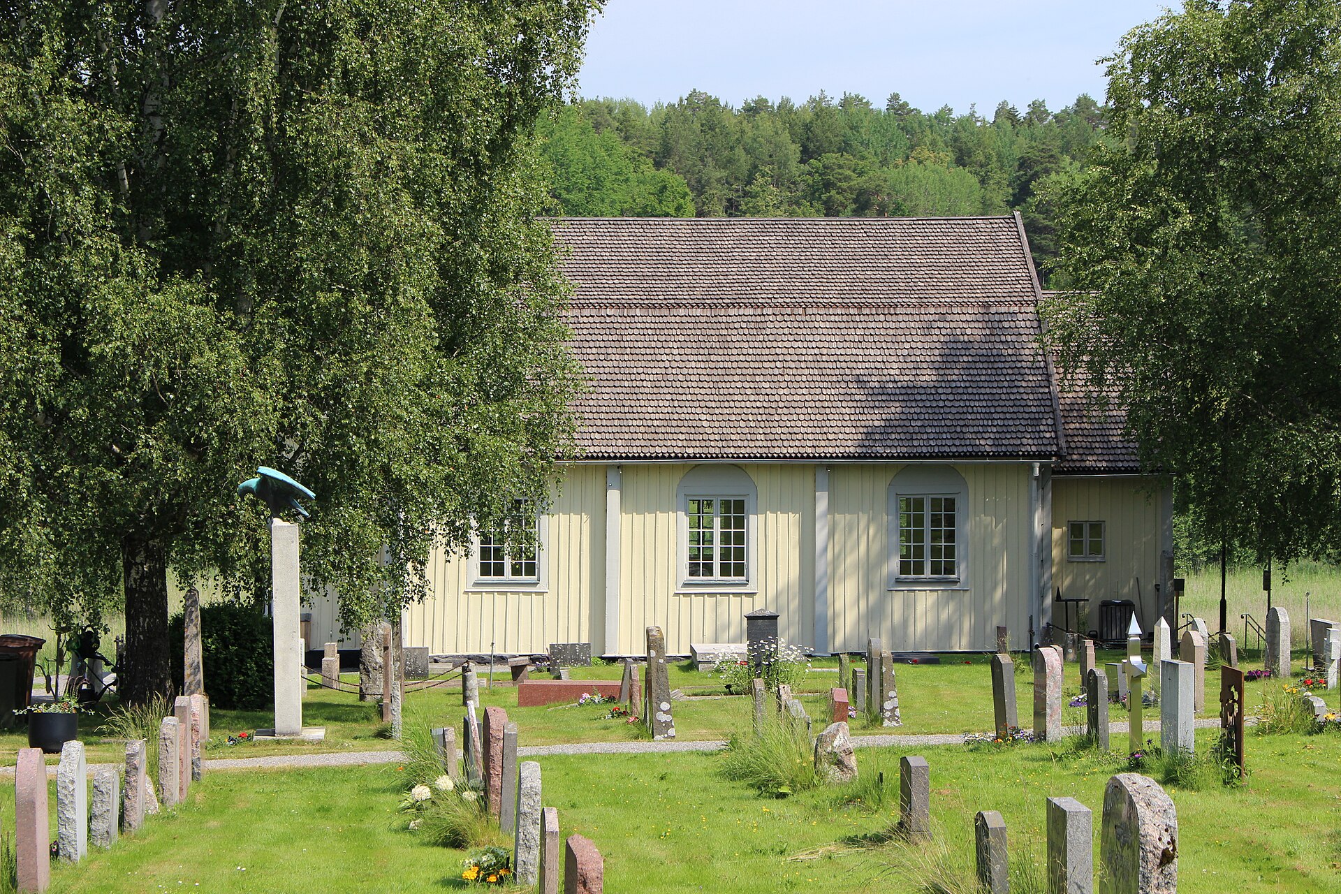 Ingarö kyrka, en ljus träkyrka med stående panel och spåntak, sedd från kyrkogården med gravstenar och en örnkrönt gravvård i förgrunden