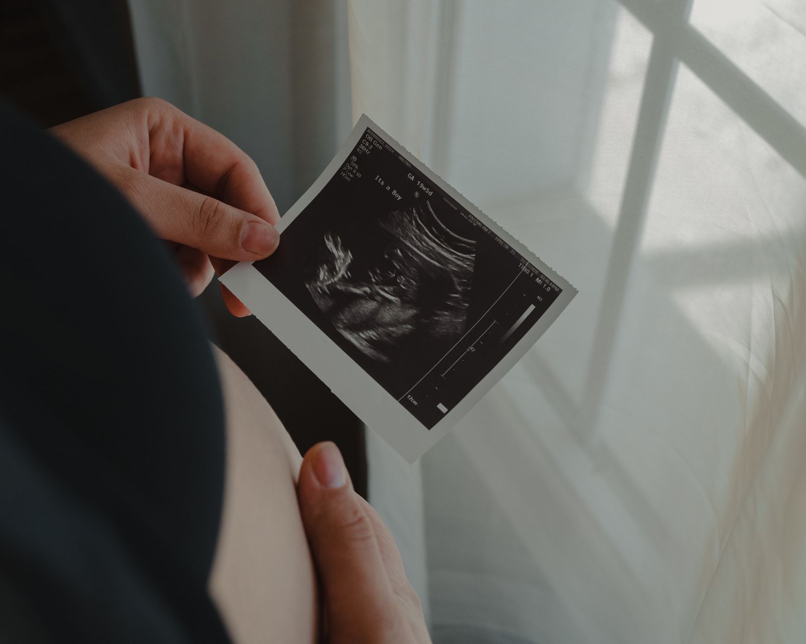 Gravid person som håller en utskriven ultraljudsbild framför magen i mjukt fönsterljus