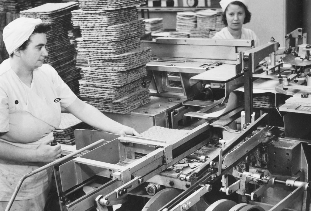 Två kvinnor i vita arbetskläder vid ett löpande band i Spisbrödsfabriken på Kvarnholmen på 1940-talet, med höga staplar av spisbröd bakom sig
