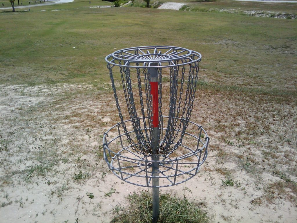 Grå discgolfkorg med hängande kedjor ovanför en trådkorg, uppställd på en öppen gräsyta