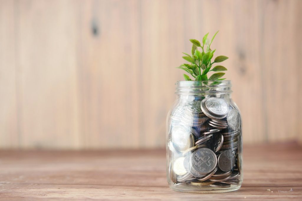 Glasburk fylld med mynt och en grön planta som växer upp ur burken, på ett träbord