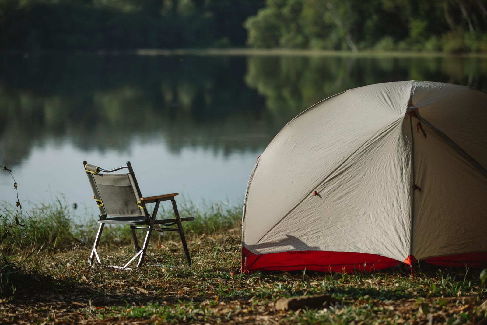 Hopfällbar campingstol med rörram står i gräset intill ett tält vid en spegelblank sjö