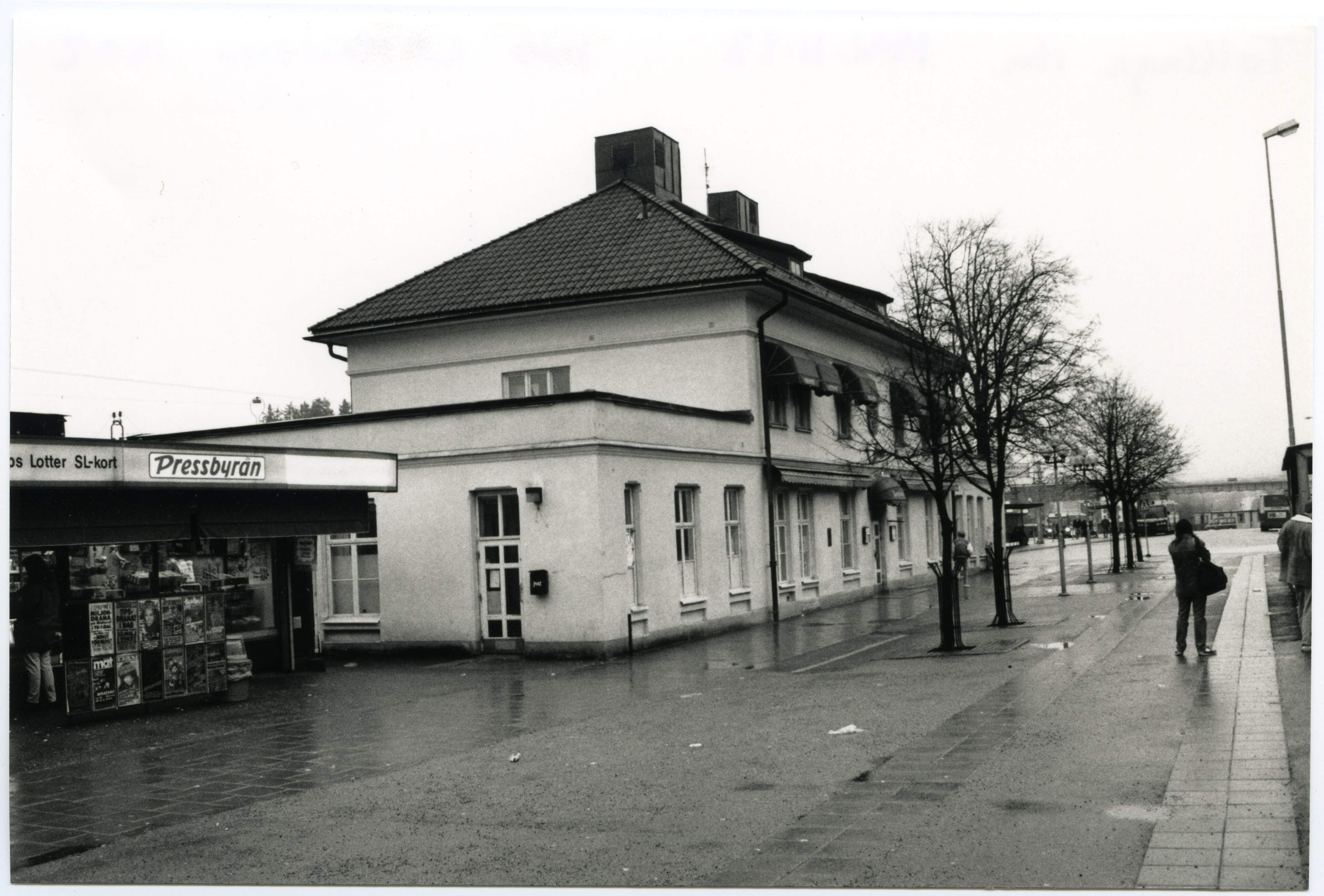 Tumba gamla stationshus i november 1991, en putsad byggnad med tegeltak, med en Pressbyråkiosk och regnvått torg framför