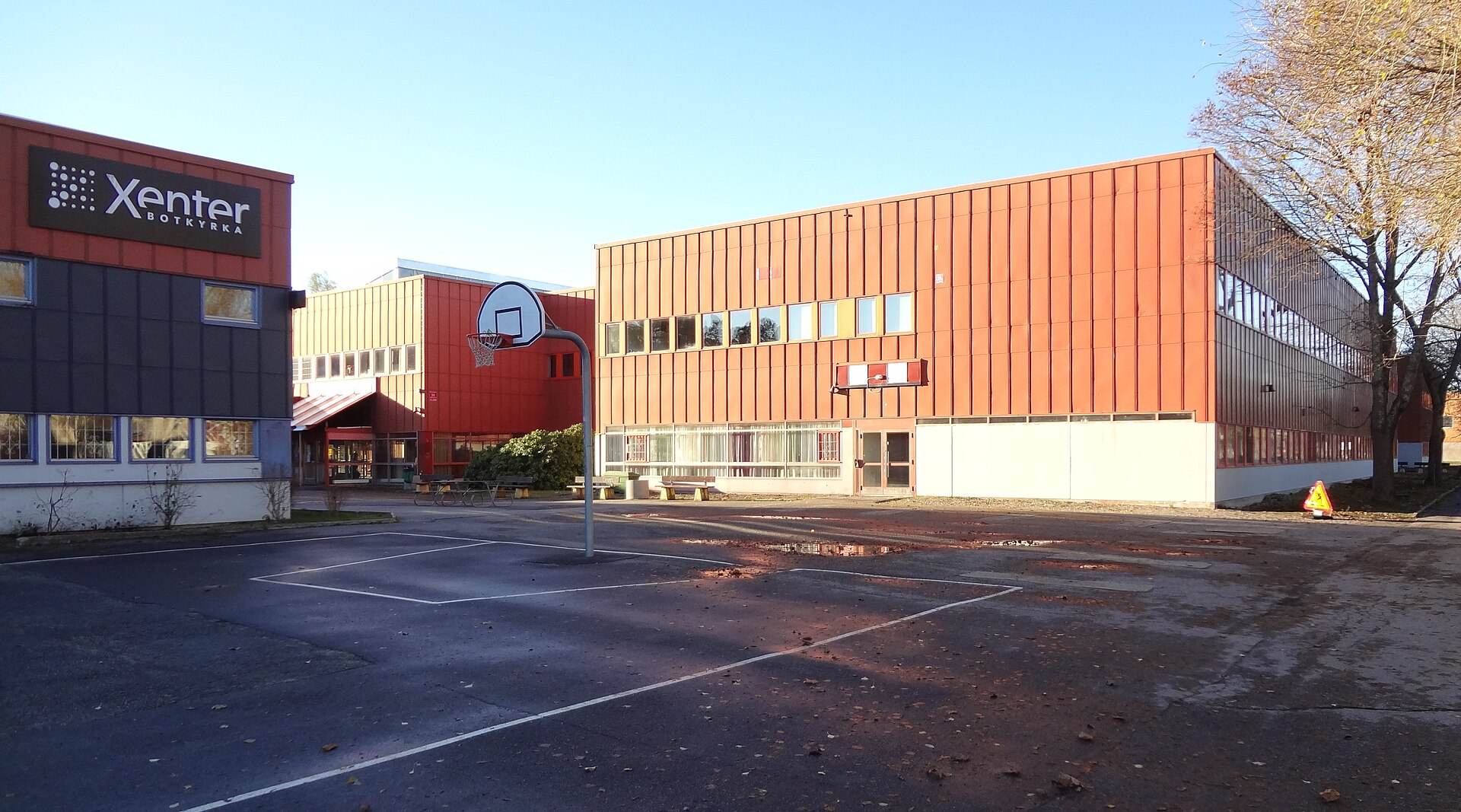 Skolgården vid Tumba gymnasium med basketkorg och röda skolbyggnader, och Xenter Botkyrka till vänster.