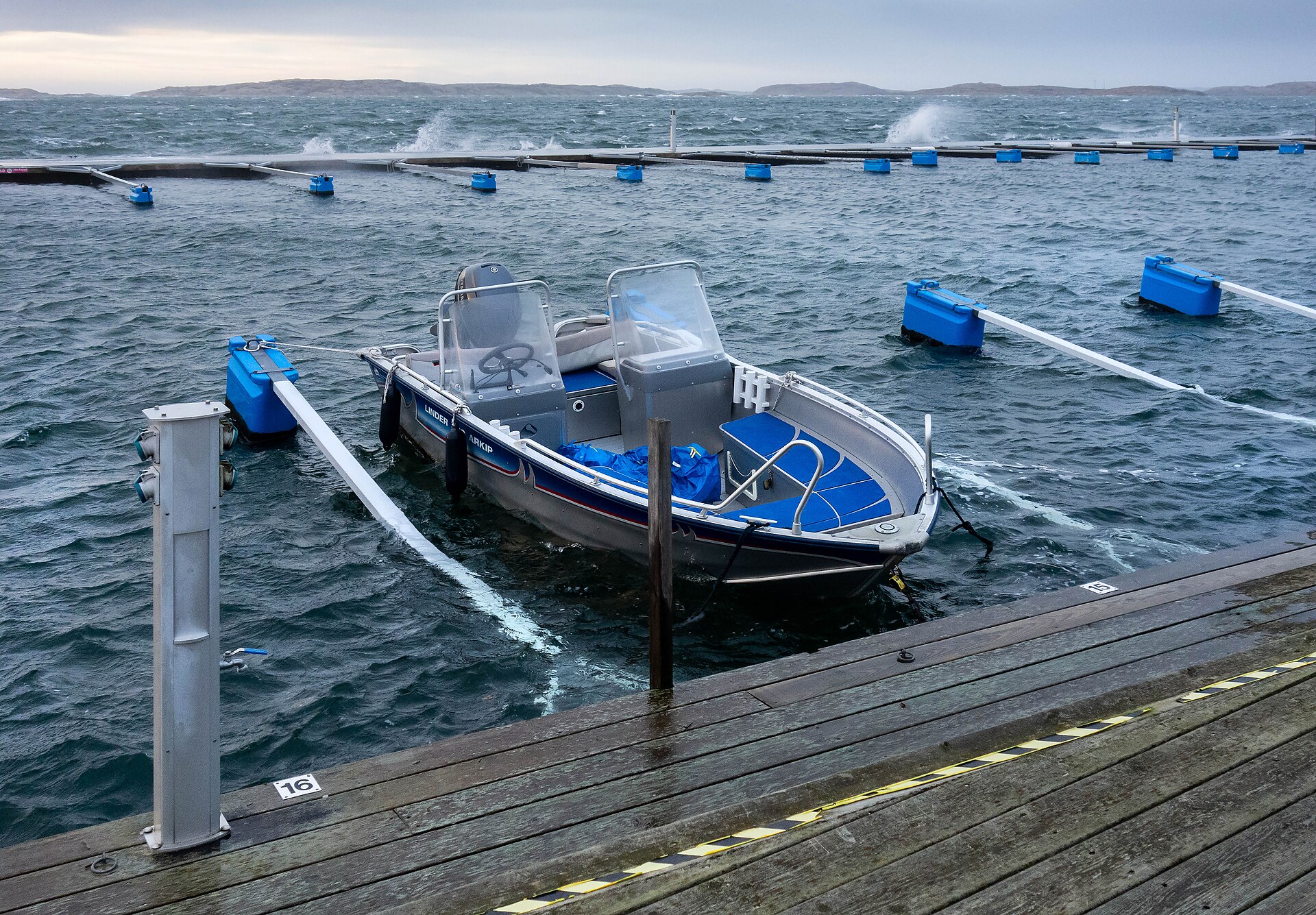 Liten öppen motorbåt förtöjd vid en brygga i Lysekil medan hård vind river upp vågor och skum över flytbryggorna bakom.