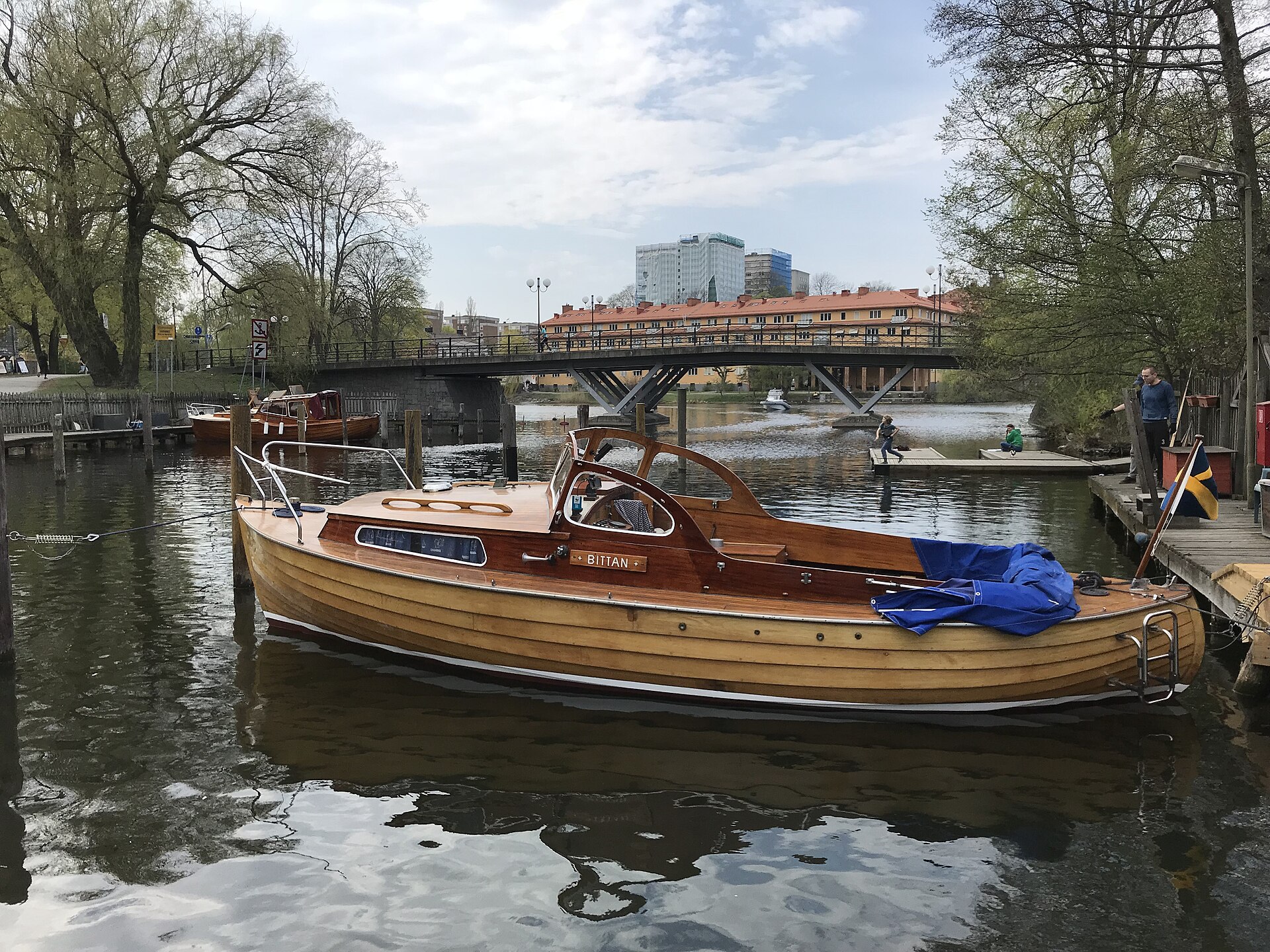 Lackad klinkbyggd träsnipa med ruff förtöjd vid en brygga i Stockholm, med en gångbro och bostadshus i bakgrunden